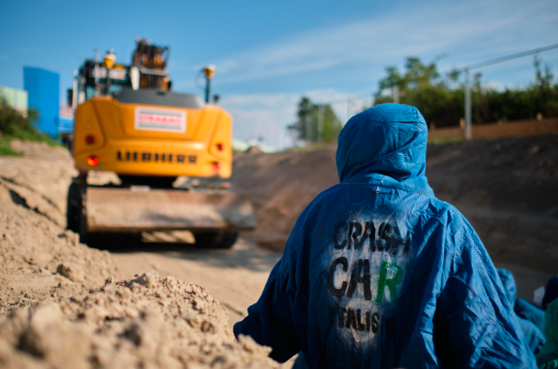 Person mit Rücken zur Kamera in einem blauen Poncho und Protestspruch am Rücken steht auf einer Baustelle einem Bagger gegenüber