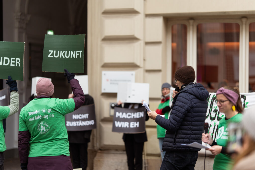Teachers for Future stehen mit Schildern wie "Kinder", "zukunft" vor dem Bildungsministerium, vor dem Personen mit Aktenordner vor dem Gesicht und Schildern wie "Machen wir eh" und "Nicht zuständig" stehen.